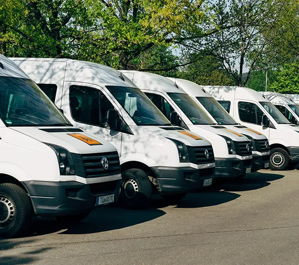 A row of white delivery vans, shielded by robust business insurance, is parked on the side of the road with trees in the background on a clear, sunny day.