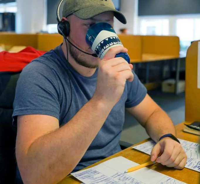 A man in a blue shirt and cap is drinking from a shark-design cup, wearing a headset, and holding a pencil at his desk in an office, likely discussing motor trade insurance with clients.