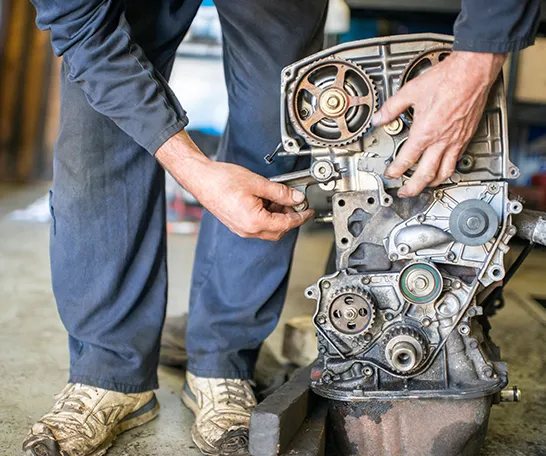 Mechanic working with their hands on an exposed engine, adjusting components. Wearing blue clothing and white sneakers, they stay focused amidst the hustle of news and views swirling around them.