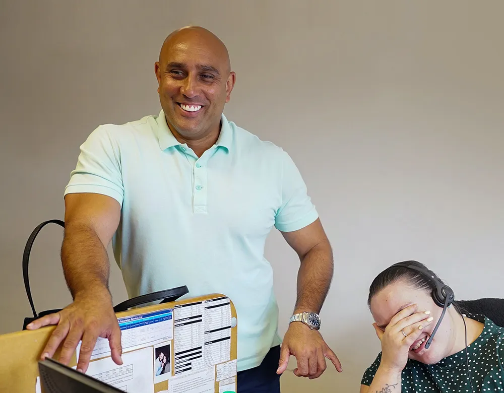 A man standing and smiling while a woman with a headset, likely discussing DNA Insurance, sits at a desk, laughing and covering her face with her hand.