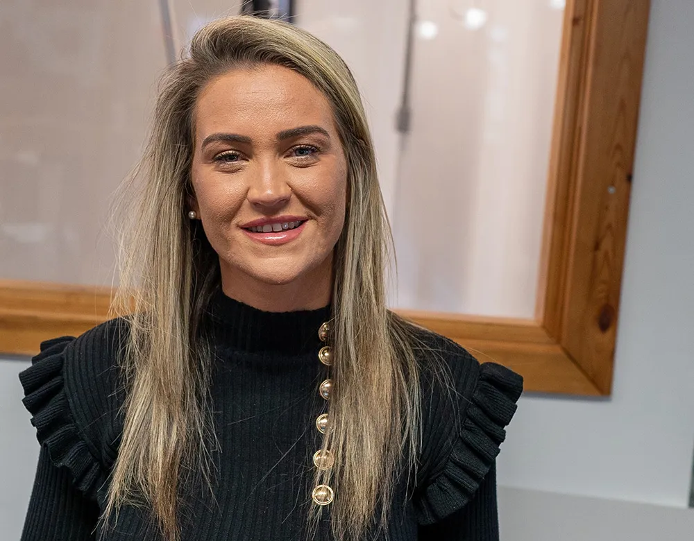 A woman with long blonde hair wearing a black sweater with gold buttons stands smiling in front of a window with a wooden frame, perhaps thinking about her recent experience getting tyre fitting insurance.