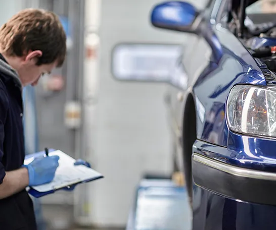 A mechanic in gloves, holding a clipboard, inspects a blue car elevated on a lift in the garage, providing keen insights for your news and views.