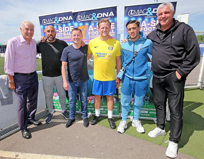 A group of six men standing together outdoors in front of event banners, dressed in casual and sports attire, smiling at the camera.