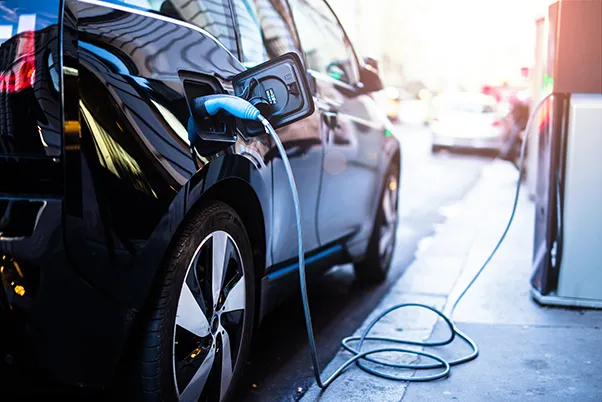 A black electric car is plugged into a charging station on a city street, with the charging cable connected to the vehicle.