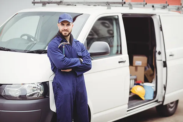 A worker in a blue jumpsuit and cap stands with crossed arms in front of a white van with an open door revealing tools and equipment inside.