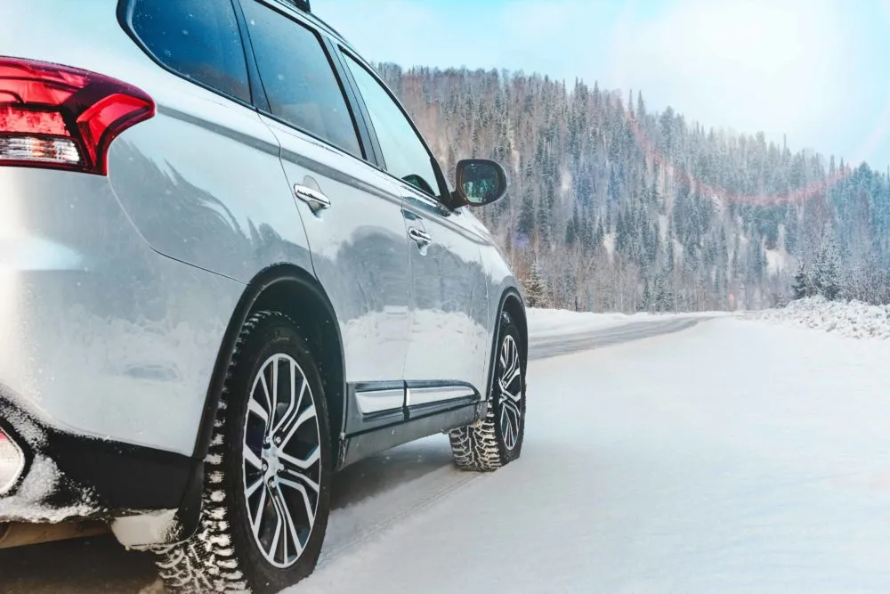 A silver SUV drives on a snowy road surrounded by frosted trees and a bright, cloudy sky.