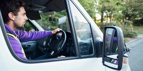 A man driving a white van looks forward, his left hand on the steering wheel. Green trees are visible outside the window.